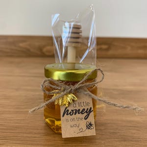 May include: A small glass jar filled with honey, tied with twine and a gold bee charm. A small wooden honey dipper is included. A brown paper tag is attached with the text "a little honey is on the way!"