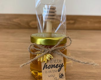 Könnte beinhalten: Ein kleines Glas gefüllt mit Honig, gebunden mit Schnur und einem goldenen Bienen-Anhänger. Ein kleiner Holz-Honiglöffel ist enthalten. Ein braunes Papieretikett ist mit dem Text "a little honey is on the way!" befestigt.