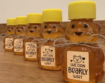 Könnte beinhalten: Reihe von Honigflaschen in Bärenform mit gelben Deckeln. Jede Flasche trägt ein Etikett mit der Aufschrift "We Can Bearly Wait" und einer Bärenillustration. Die Flaschen sind mit Bindfaden gebunden.