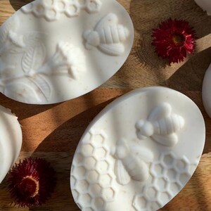 K&ouml;nnte beinhalten: Ovale, cremefarbene Seifen mit Bienen- und Wabenmustern. Die Seifen sind auf einer Holzoberfl&auml;che mit roten Blumen angeordnet. Die Seifen haben ein detailliertes, gepr&auml;gtes Design einer Biene und einer Wabe.