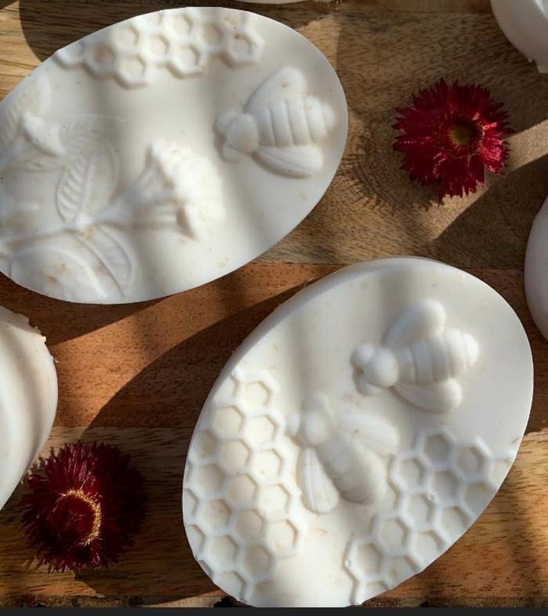 K&ouml;nnte beinhalten: Ovale, cremefarbene Seifen mit Bienen- und Wabenmustern. Die Seifen sind auf einer Holzoberfl&auml;che mit roten Blumen angeordnet. Die Seifen haben ein detailliertes, gepr&auml;gtes Design einer Biene und einer Wabe.