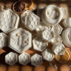 K&ouml;nnte beinhalten: Eine Sammlung wei&szlig;er Seifen mit Bienenmotiven in verschiedenen Formen, darunter Sechsecke und Kreise. Einige Seifen zeigen ein Bienendesign, andere ein Wabenmuster. Getrocknete Orangenscheiben und Haferflocken sind verstreut.