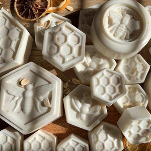 K&ouml;nnte beinhalten: Eine Sammlung handgemachter, cremefarbener Seifen mit Bienen- und Wabenmustern. Einige Seifen sind sechseckig, andere rund. Getrocknete Orangenscheiben und Haferflocken sind um die Seifen verstreut.