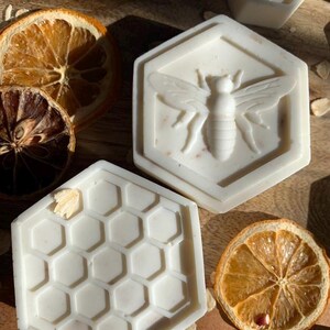 K&ouml;nnte beinhalten: Sechseckige, cremefarbene Seifen mit Bienen- und Wabenmustern. Die Seife mit der Biene hat ein detailliertes Relief des Insekts. Getrocknete Orangenscheiben und Haferflocken sind um die Seifen herum verstreut.
