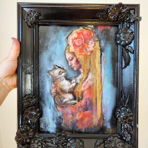 May include: An oil painting in a black ornate frame. The artwork depicts a woman with long blonde hair and a floral headpiece holding a cat. The woman is wearing a red and orange dress. The frame has decorative floral accents.