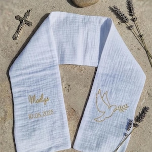 May include: White baptismal stole with gold embroidered dove holding an olive branch. The stole has the name "Maelys" and the date "10.05.2025" embroidered in gold. A small stone with a dove carving, a silver cross, and lavender sprigs are also present.