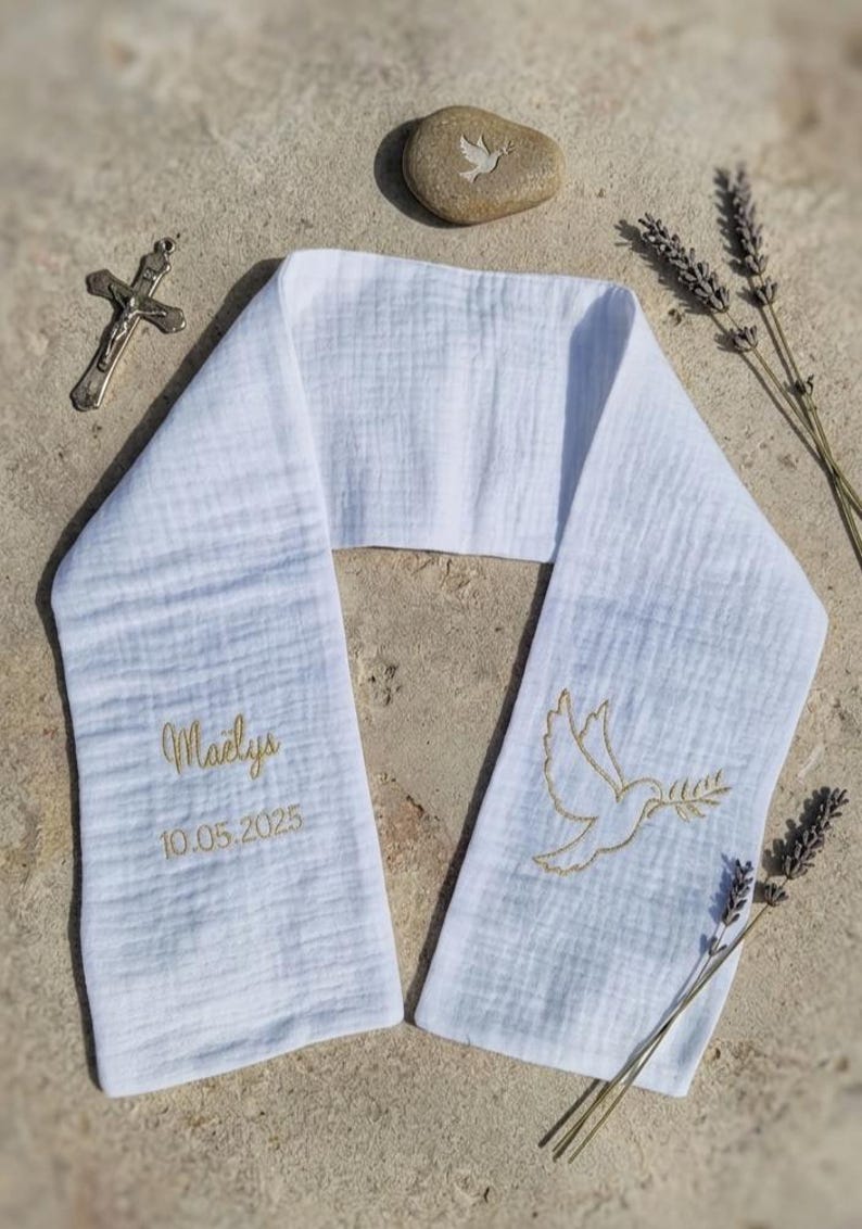 May include: White baptismal stole with gold embroidered dove holding an olive branch. The stole has the name "Maelys" and the date "10.05.2025" embroidered in gold. A small stone with a dove carving, a silver cross, and lavender sprigs are also present.
