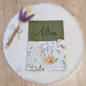 Peut inclure: Un livre en tissu vert et blanc avec le nom "Albin" brodé sur la couverture. Le livre présente un renard blanc et un motif floral.