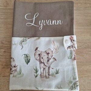 Peut inclure: Une couverture de livre en tissu marron avec le nom "Lyann" brodé sur le devant. Le dos de la couverture est en tissu blanc avec un motif répété d'éléphants, d'oiseaux et de feuilles.