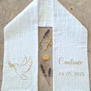 Peut inclure: Étole de baptême blanche avec broderie dorée d'une colombe tenant un rameau d'olivier. L'étole porte le nom "Constance" et la date 04.05.2025 brodés en or. Une croix et un médaillon dorés sont également présents.