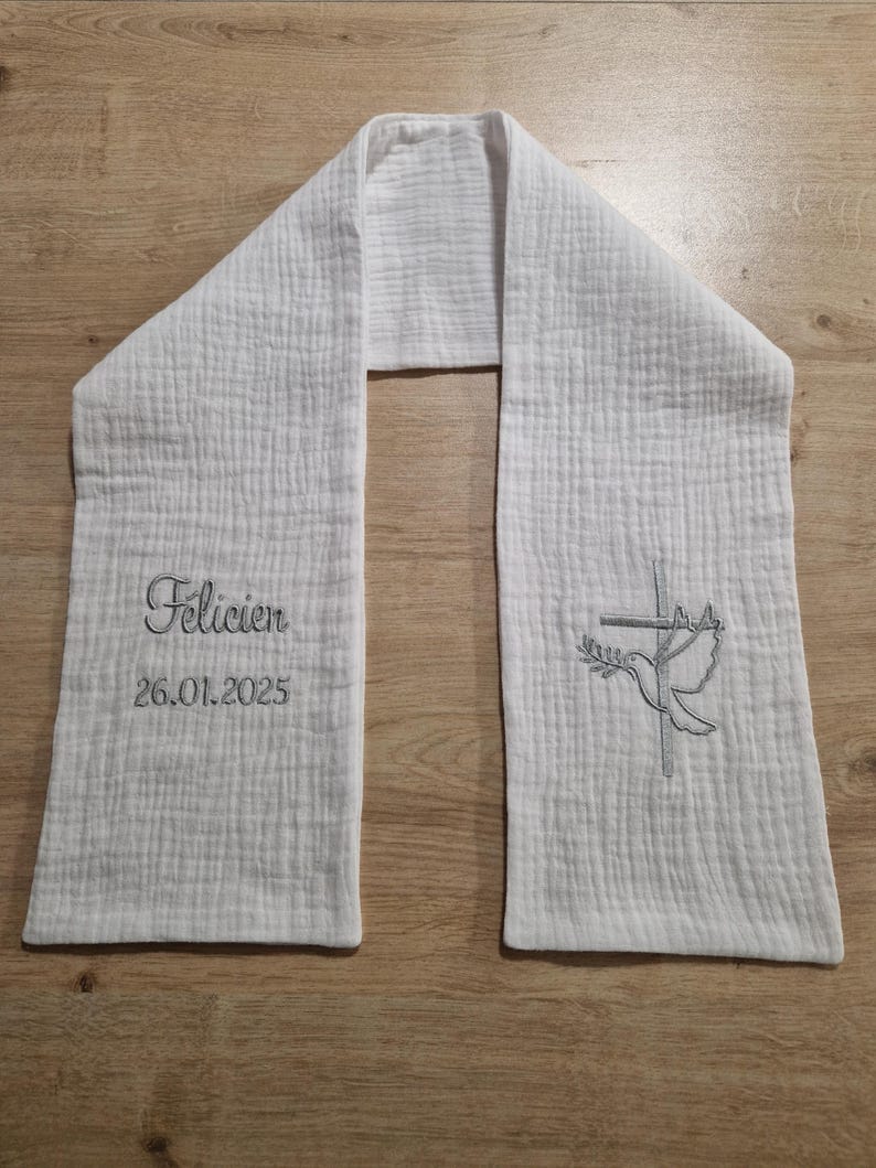 May include: White baptismal stole with embroidered details. The stole features the name "Felicien" and the date "26.01.2025" in script. A dove and cross design is embroidered on the opposite end. The stole is made of a textured fabric.