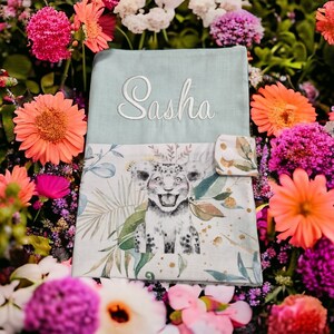 Peut inclure: Un livre pour bébé bleu clair avec un nom brodé en blanc "Sasha" sur le devant. Le livre a une poche sur le devant et une illustration colorée d'un léopard blanc au dos.