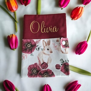 Peut inclure: Une couverture de livre en tissu bordeaux avec une fermeture à bouton blanc. La couverture avant présente un motif floral avec un lapin blanc et le nom "Olivia" en lettres dorées.