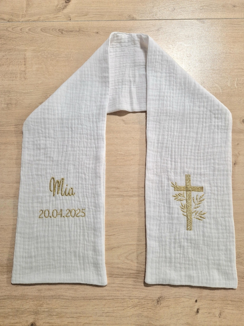 May include: White baptismal stole with a textured surface. The stole features gold embroidered text that reads "Mia" and "20.04.2025". A gold embroidered cross with floral accents is also present. The stole is made of a soft, lightweight fabric.