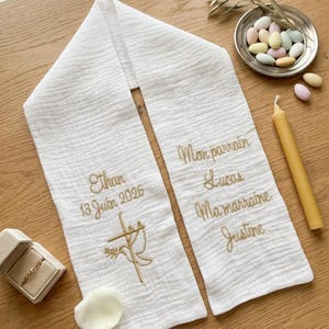 May include: White baptismal stole with gold embroidery. The stole features the name "Ethan" and the date "13 Juin 2026", along with a cross and dove design. The other side has names "Lucas" and "Justine".