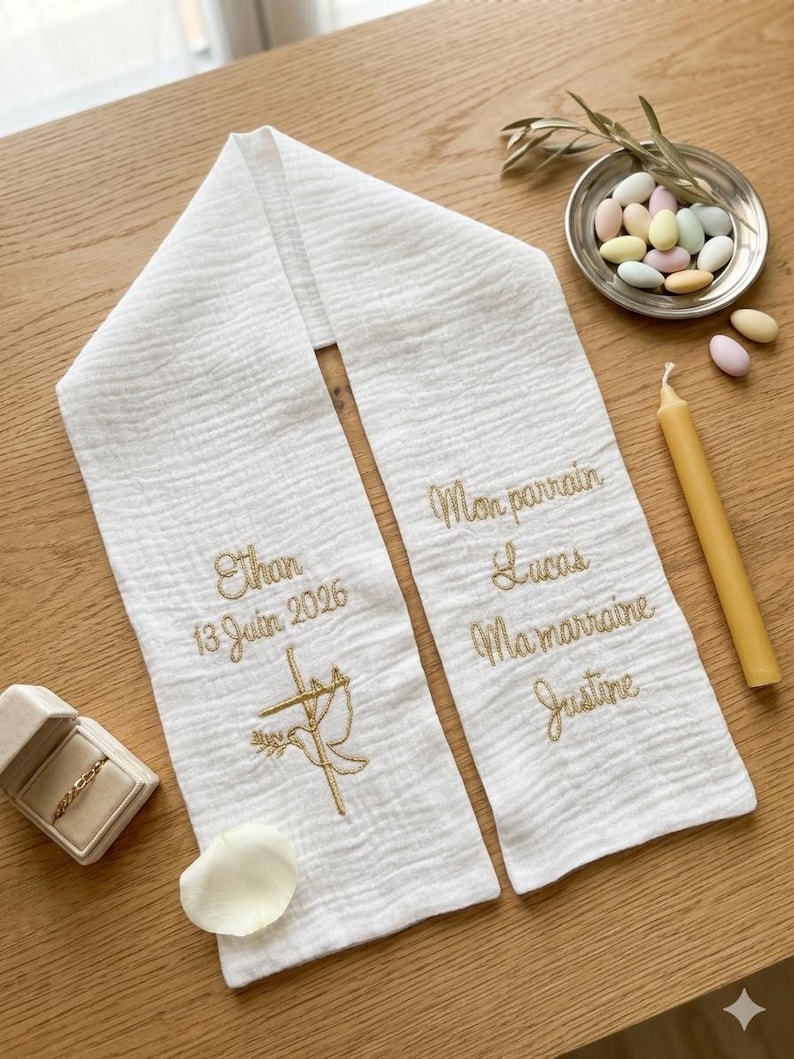 May include: White baptismal stole with gold embroidery. The stole features the name "Ethan" and the date "13 Juin 2026", along with a cross and dove design. The other side has names "Lucas" and "Justine".