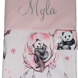 Peut inclure: Un livre pour bébé en tissu rose avec un rabat blanc. Le rabat a un bouton blanc et un motif d'ours panda. Le devant du livre est rose et porte le nom "Myla" écrit en lettres argentées. Le dos du livre a un fond blanc avec un ours panda assis sur une branche avec des fleurs roses. L'ours panda est entouré de plumes.