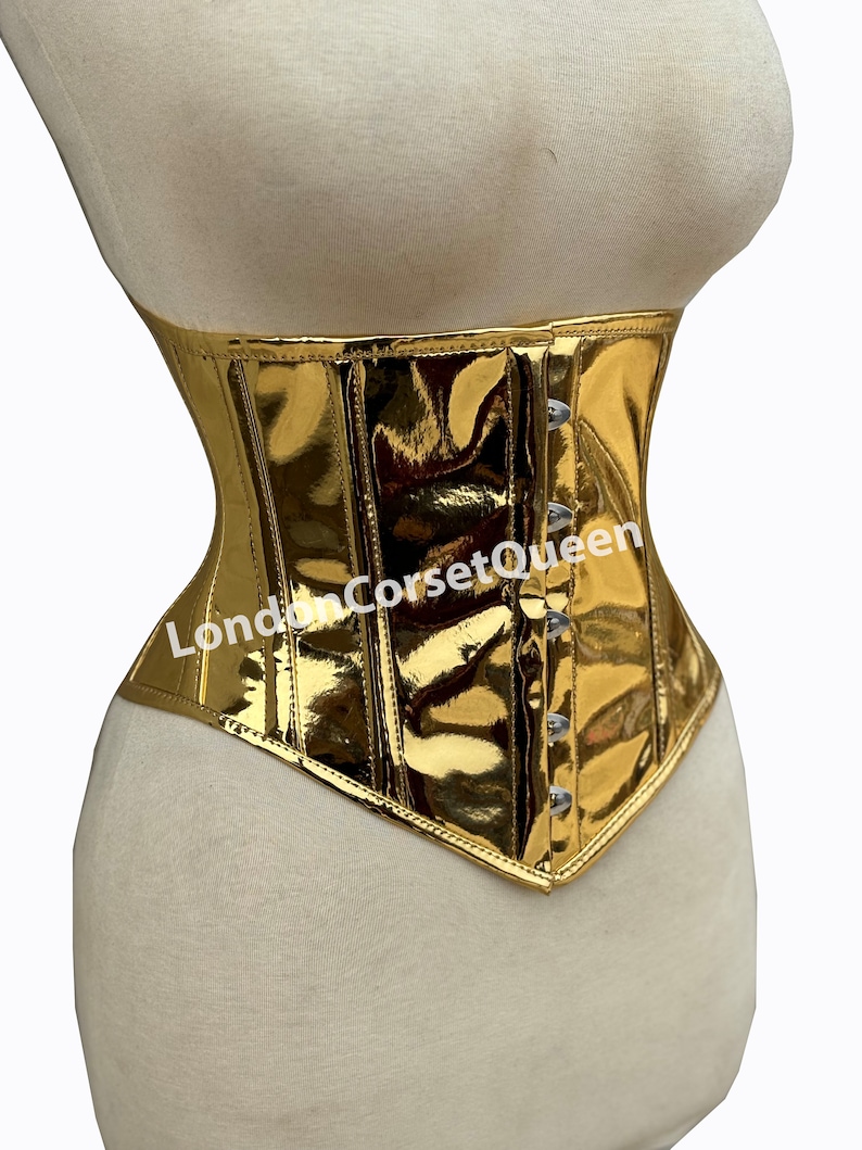 Puede incluir: Un cors&eacute; met&aacute;lico dorado con la parte inferior puntiaguda y cierre de busk. El cors&eacute; tiene un acabado brillante y est&aacute; hecho de una tela suave. El texto "LondonCorsetQueen" es visible en la parte delantera del cors&eacute;.