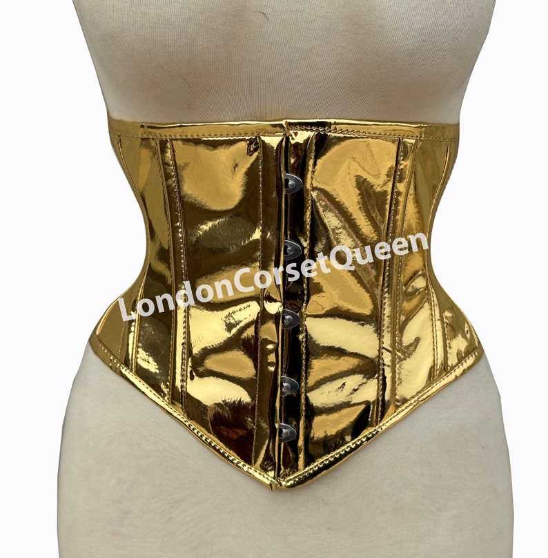 Puede incluir: Un cors&eacute; met&aacute;lico dorado con la parte inferior puntiaguda y cierre frontal con ganchos met&aacute;licos. El cors&eacute; tiene un acabado brillante y reflectante y est&aacute; etiquetado como "LondonCorsetQueen".