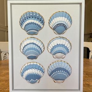 May include: A rectangular white frame containing six scallop shells arranged in two columns. Each shell is painted with varying patterns of light blue, dark blue, and white, with gold-painted edges. The shells are mounted on a plain white background.