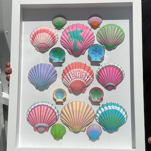 May include: A framed artwork featuring a collection of colorful, painted seashells. The shells are in various sizes and colors, including pink, green, blue, and yellow. Some shells have the word "BEACH" painted on them, and others feature palm tree designs. The frame is white.