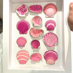 May include: A white framed shadow box featuring a collection of painted seashell art. The shells are in shades of pink, with gold trim and various patterns. The arrangement includes different shell shapes and sizes.