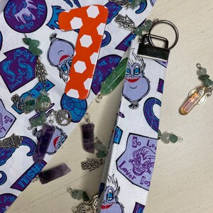 Pu&ograve; includere: Un portachiavi in tessuto bianco con un'immagine di Ursula de La Sirenetta in viola e blu. Il portachiavi ha il testo "Lover Bos" e "So Lo" su di esso. Il portachiavi &egrave; attaccato a un portachiavi argentato.