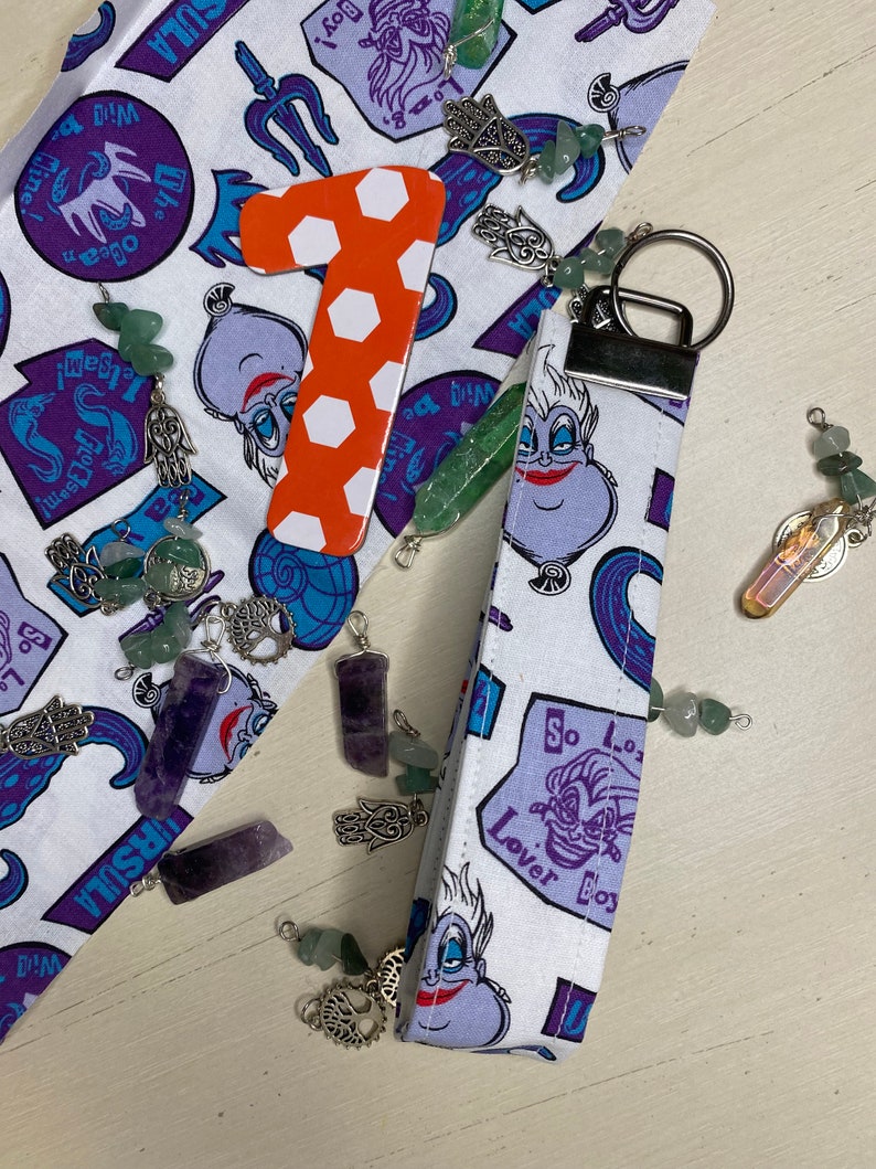 Pu&ograve; includere: Un portachiavi in tessuto bianco con un'immagine di Ursula de La Sirenetta in viola e blu. Il portachiavi ha il testo "Lover Bos" e "So Lo" su di esso. Il portachiavi &egrave; attaccato a un portachiavi argentato.
