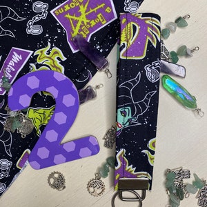 Pu&ograve; includere: Portachiavi in tessuto viola e nero con un design viola e giallo che presenta un'immagine di cartone animato di Malefica dal film Disney La Bella Addormentata nel Bosco. Il portachiavi ha un anello portachiavi argentato.