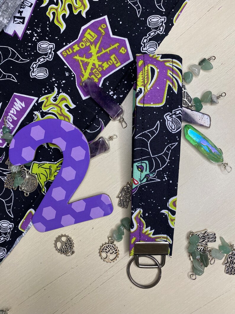 Pu&ograve; includere: Portachiavi in tessuto viola e nero con un design viola e giallo che presenta un'immagine di cartone animato di Malefica dal film Disney La Bella Addormentata nel Bosco. Il portachiavi ha un anello portachiavi argentato.