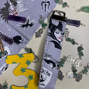 Pu&ograve; includere: Un portachiavi in tessuto grigio e nero con un'immagine di cartone animato di Malefica de La Bella Addormentata nel Bosco di Disney. Il portachiavi presenta un anello portachiavi argentato e un ciondolo argentato con un design ad albero. Il portachiavi &egrave; circondato da pietre verdi e viola e ciondoli argentati.