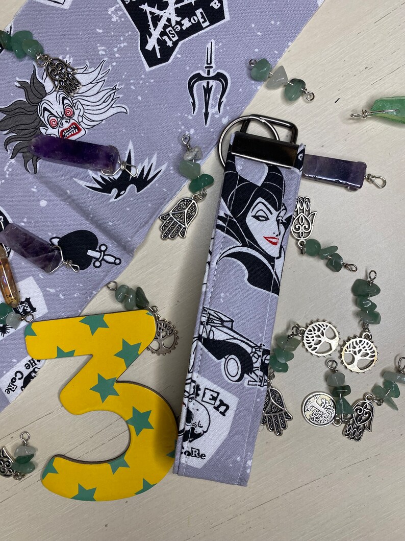 Pu&ograve; includere: Un portachiavi in tessuto grigio e nero con un'immagine di cartone animato di Malefica de La Bella Addormentata nel Bosco di Disney. Il portachiavi presenta un anello portachiavi argentato e un ciondolo argentato con un design ad albero. Il portachiavi &egrave; circondato da pietre verdi e viola e ciondoli argentati.