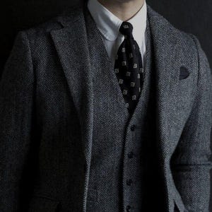 May include: A grey herringbone suit with a black waistcoat and a black tie with a white geometric pattern.