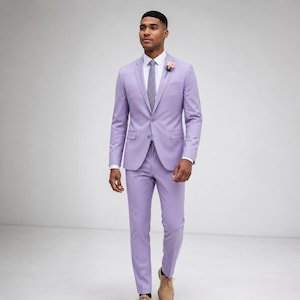 May include: A lilac suit comprising a jacket and trousers. The jacket features a two-button fastening and a flower in the lapel. The suit is worn with a white shirt and a grey tie. The person is wearing brown shoes.