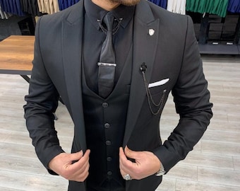 May include: A black three-piece suit with a matching black shirt and tie. The suit features a blazer, vest, and trousers. The tie is secured with a tie clip, and a small chain with a flower detail is attached to the lapel. Black leather shoes complete the ensemble.