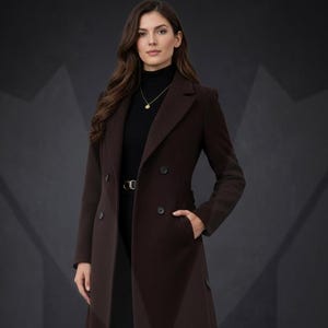 May include: A long, dark brown coat with a double-breasted design. The coat features a notched collar, two rows of buttons, and a belt at the waist. The model is wearing a black turtleneck and black pants. The coat has pockets.