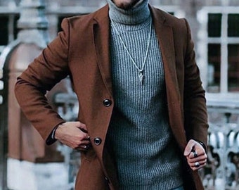 May include: A brown overcoat with two buttons, worn over a gray turtleneck sweater and blue jeans. The outfit is accessorized with a black hat and a silver necklace. The coat has a classic, tailored design.