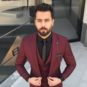 May include: A three-piece burgundy suit with a black shirt and tie. The suit includes a blazer, waistcoat, and trousers. The outfit is completed with black loafers. A silver chain is attached to the suit pocket.