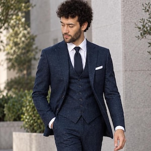 May include: A three-piece navy blue suit with a subtle speckled texture, consisting of a single-breasted blazer, a matching button-up vest, and trousers. The outfit includes a crisp white collared shirt, a dark navy tie, and brown leather shoes.
