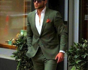 Puede incluir: Un traje verde oliva con una camisa blanca y un pañuelo de bolsillo rojo. El traje es de color liso y se lleva con botas de cuero marrones. La persona lleva gafas de sol negras.