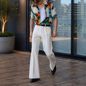 May include: A person wearing a colorful short-sleeve button-down shirt, white undershirt, and cream-colored flared pants. The shirt has a vibrant pattern with orange, blue, and green hues. Black belt and shoes complete the outfit.