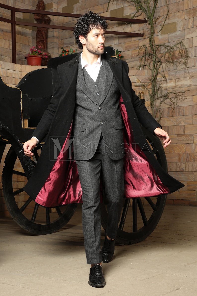 May include: A black overcoat with a red lining and a grey suit underneath. The coat is open and the lining is visible. The person is standing in front of a wooden carriage wheel.