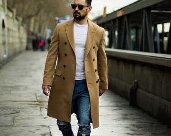 May include: A man wearing a camel-colored double-breasted coat, a white t-shirt, blue jeans with distressed details, and white sneakers. He is walking on a paved sidewalk.