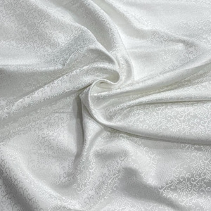 May include: White fabric with a subtle floral pattern. The fabric is draped, creating soft folds and shadows. The pattern is visible across the surface, adding texture and visual interest. This fabric is suitable for apparel or home decor.
