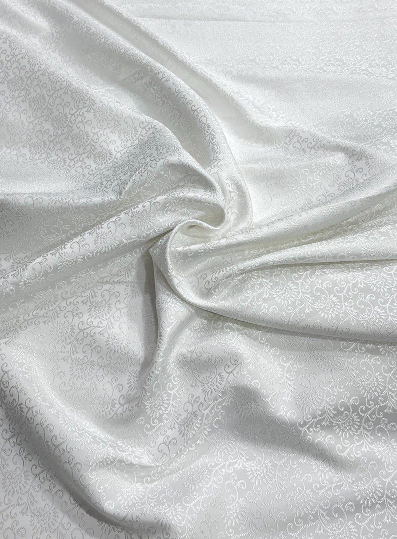 May include: White fabric with a subtle floral pattern. The fabric is draped, creating soft folds and shadows. The pattern is visible across the surface, adding texture and visual interest. This fabric is suitable for apparel or home decor.