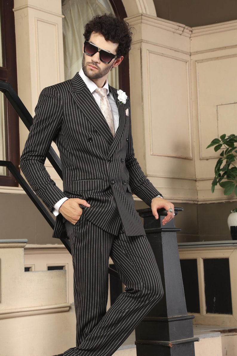 May include: A double-breasted suit in black with thin white vertical pinstripes, featuring a white collared shirt, a light-colored patterned necktie, a white lapel flower, and dark-tinted sunglasses, worn with matching pinstriped trousers.