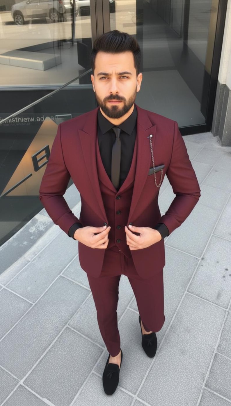 May include: A three-piece burgundy suit with a black shirt and tie. The suit includes a blazer, waistcoat, and trousers. The outfit is completed with black loafers. A silver chain is attached to the suit pocket.