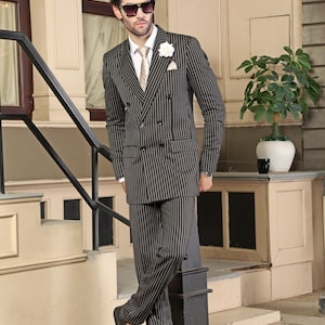 May include: A dark charcoal double-breasted suit with thin white pinstripes, featuring a white dress shirt, a patterned beige necktie, a white lapel flower, a pocket square, black sunglasses, and black leather dress shoes.