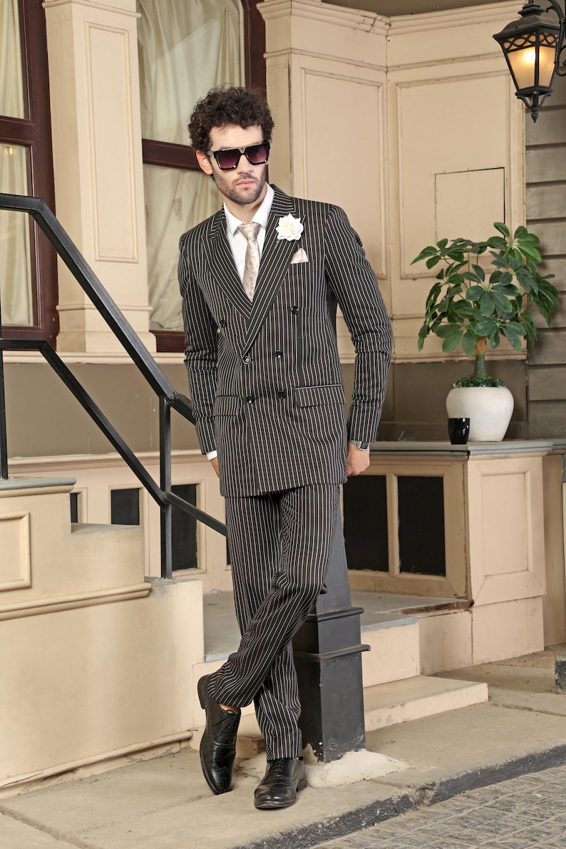 May include: A dark charcoal double-breasted suit with thin white pinstripes, featuring a white dress shirt, a patterned beige necktie, a white lapel flower, a pocket square, black sunglasses, and black leather dress shoes.