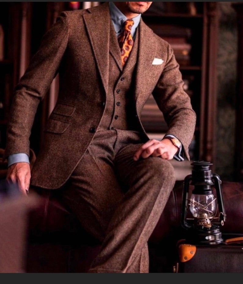 May include: A brown tweed three-piece suit with a waistcoat and a patterned tie. The suit is being worn by a person sitting on a brown leather chair with a lantern on a suitcase beside them.